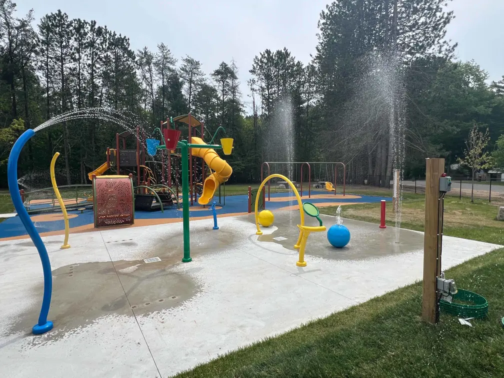 Water sprays from multiple colourful splash pad features on a concrete pad next to a playground at the Lake of Bays Community Centre & Arena in Baysville