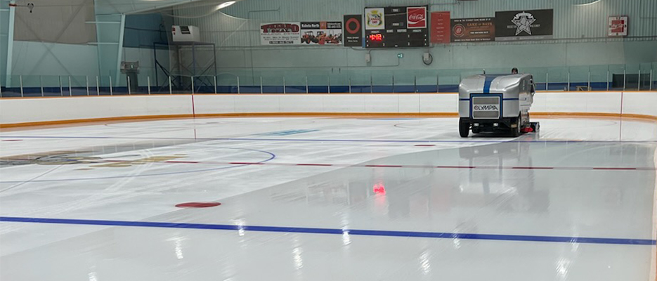 Indoor ice rink at the Lake of Bays arena freshly resurfaced by a blue and white Zamboni machine, with painted lines and circles on the ice.