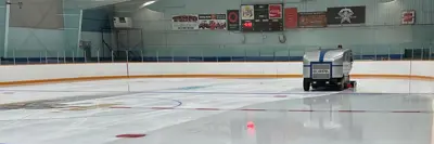 Indoor ice rink at the Lake of Bays arena freshly resurfaced by a blue and white Zamboni machine, with painted lines and circles on the ice.