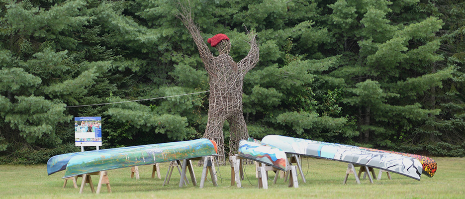 A 19-foot-tall arboreal sculpture situated next to seven canoes with murals painted on them.