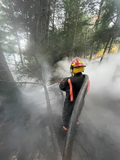Firefighter surrounded by thick smoke, spraying water on a burning patch.